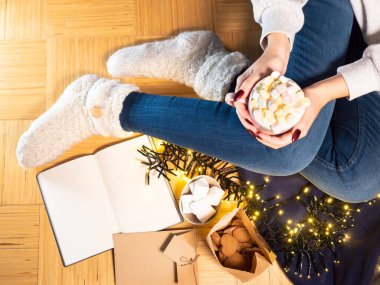 Kadın elleri elinde bir fincan sıcak çikolata, marşmelov ve örülmüş çoraplar, yerde bir kitap.