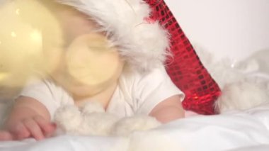 6 aylık Noel Baba şapkalı güzel bir kızın portresi beyaz yatakta uzanıyor ve oyuncaklarla oynuyor. Mutlu Noeller ve mutlu yıllar.