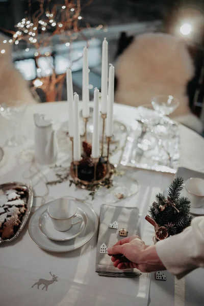 Woman hand setting up table for Christmas dinner. Table is decorated with white tablecloth, cups, glasses, candlestick, table-napkin and small wooden decorative houses