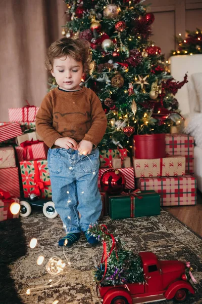 Очаровательный Кудрявый Мальчик Играет Рождественскими Огнями Уютной Рождественской Украшенной Комнате — стоковое фото
