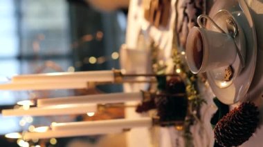 View of christmas table setting in white and golden colors at evening. Close-up view of cup of hot tea with steam, burning candles. Christmas eve. Vertical video