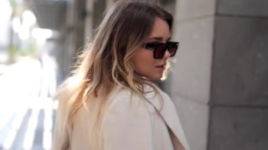 Modern şehir sokaklarında poz veren beyaz ceketli güzel bir kadın. Kameraya bakıyor, poz veriyor, etrafında dönüyor.