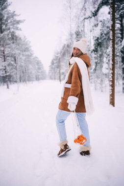 Sıcak yün ceketli, beyaz atkılı, şapkalı ve eldivenli, mandalina çantasıyla poz veren, karlı kış ormanlarında yürüyen mutlu ve çekici genç bir kadın.