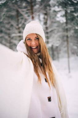 Active beautiful young woman in warm white sheepskin coat, hat and mittens and flying scarf enjoying snowy winter forest, looking ahead