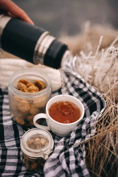 Female hand pouring hot tea into white cup from thermos at snowy winter nature. Winter picnic near the sea or lake, travel, adventure concept. 