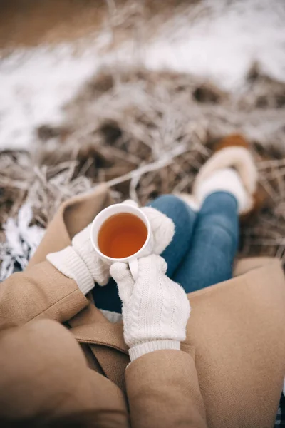 Unrecognizable woman in white woolen mittens holding cup of tea enjoying winter frozen nature. Morning breakfast at nature. Travel, adventure concept. 