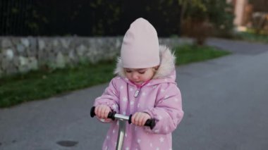 Adorable toddler girl in spring or autumn overall riding a childrens scooter on the street 