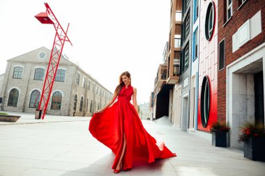 Young charming woman in long flying red dress and red shoes walking down the modern new street 