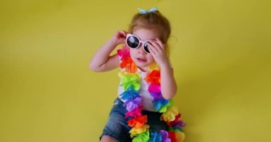 Adorable little girl in white t-shirt wearing summer sunglasses sitting on yellow background, studio portrait. Summer vacations