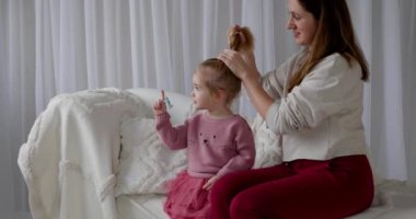 Loving mother sit on white sofa at home and combs hair of adorable 2 years old daughter. 