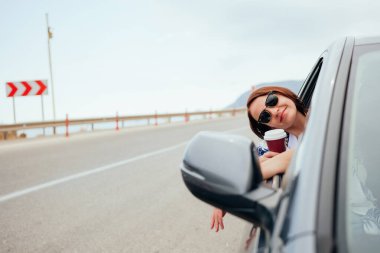 Elinde hazır kahveyle pencereden dışarı bakan mutlu gülümseyen genç bayan yolcu. Keyifli araba yolculuğu, yaz tatili.