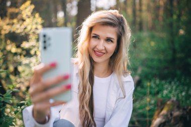 Sarışın, uzun saçlı, gülümseyen genç bir kadın ormanda cep telefonu kullanarak selfie çekiyor. Dışarıda yürürken arkadaşınla konuş.