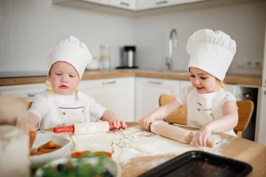 Sevimli 3 yaşındaki erkek ve kız aşçı şapkalı ve önlüklü mutfakta hamur işi yapıyor. Genç yardımcılar, lezzetli hamur işleri hazırlıyorlar..