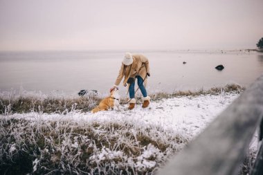 Happy attractive young woman with white scarf, hat and mittens playing with her welsh corgi pembroke puppy outdoors by the sea, snowy winter field. 