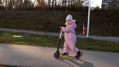 Adorable toddler girl in spring or autumn overall riding a childrens scooter on the street 