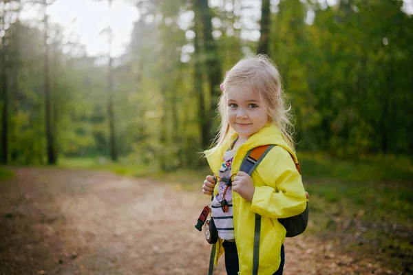 Mutlu anaokulu kızı sonbahar ormanı başlarında sırt çantasıyla kameraya bakarak yürüyor. Çocuklarla açık hava aktiviteleri
