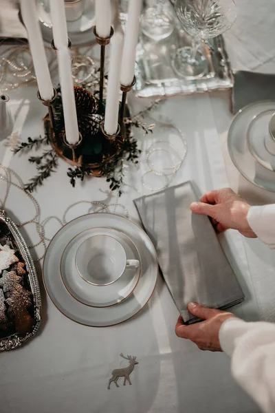 Woman hand setting up table for Christmas dinner. Table is decorated with white tablecloth, cups, ginger bread cookies, candlestick, table napkin and small wooden decorative houses