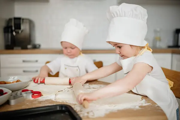 Adorable 3 years old boy and girl in chef hats and aprons rolling out pastry dough at the kitchen. Young helpers, preparing delicious pastry food.