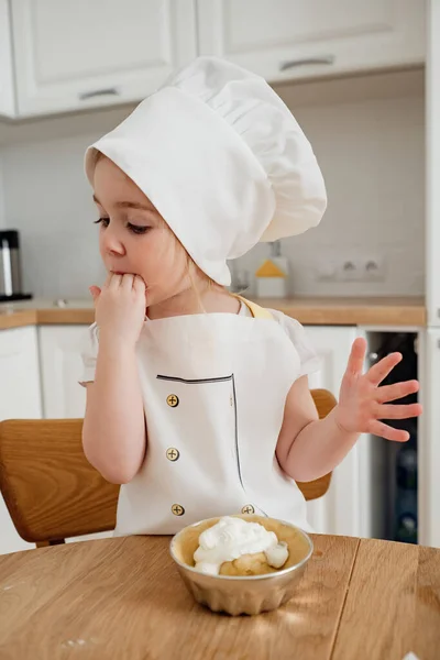 Adorable girl in chef hat and aprons preparing cake at the kitchen. Child licking fingers. Delicious sweet food.