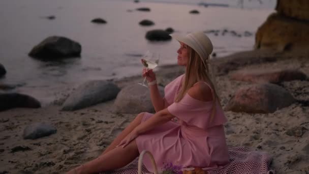 イチゴの帽子とピンクのドレスに座っている若い女性は 海の日の出の砂浜でワインとピクニックを持っています 屋外ロマンチックな夏の夜 — ストック動画