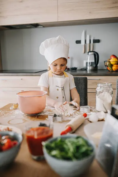 Adorable girl in chefs hats and aprons playing with the flour in the kitchen, preparing pizza. Kids hobbies