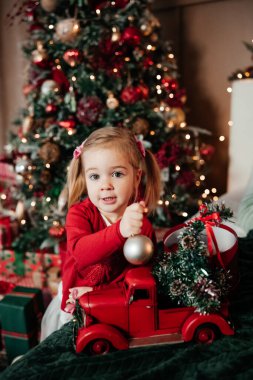 İki yaşında, kırmızı kazaklı tatlı bir kız Noel arabasıyla oynuyor. Sıcak Noel evi