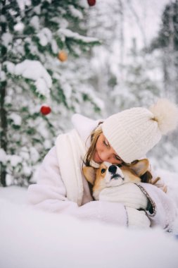 Beyaz atkısı, şapkası ve eldivenleri olan mutlu ve çekici genç bir kadın, karlı kış ormanlarında Noel süslenmiş köknar ağacının altında oturmuş Galli Corgi Pembroke köpeğine sarılıyor.. 