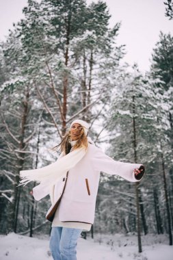 Aktif, beyaz yün ceketli, şapkalı ve uçan atkılı güzel bir kadın karlı kış ormanının tadını çıkarıyor.