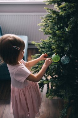Mercan pembesi elbiseli, Noel ağacını taşaklarla süsleyen iki yaşında sevimli bir kız çocuğu. Sıcak Noel evi