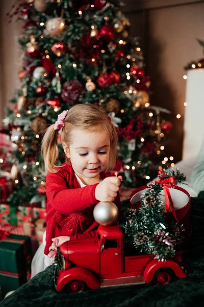 İki yaşında, kırmızı kazaklı tatlı bir kız Noel arabasıyla oynuyor. Sıcak Noel evi