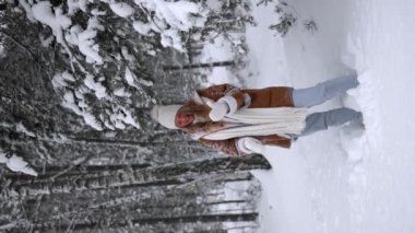 Young beautiful woman in warm sheepskin coat, hat and mittens throw snow up walking in snowy winter forest. Vertical video