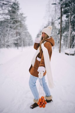 Sıcak yün ceketli, beyaz atkılı, şapkalı ve eldivenli, mandalina çantasıyla poz veren, karlı kış ormanlarında yürüyen mutlu ve çekici genç bir kadın.