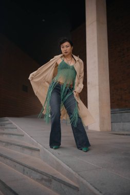 Bir bayan dansçı, bej bir ceket giyiyor ve moda süveteri ile şehirde şık adımlarla poz veriyor.