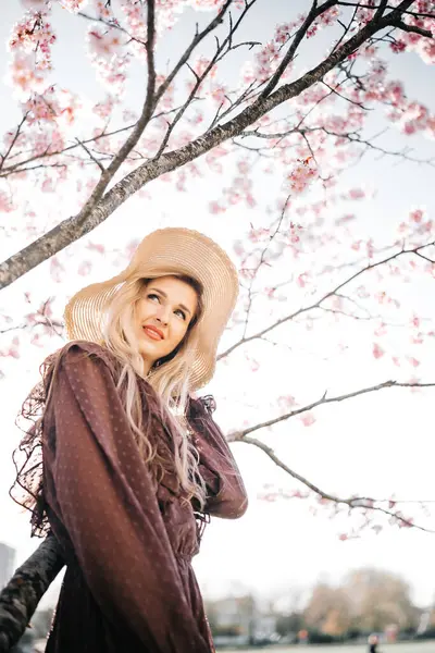 若いセンセーショナルかわいいブロンドの女性の肖像画 花開く桜の木に立っている帽子 ロイヤリティフリーのストック写真