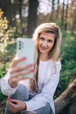 Sarışın, uzun saçlı, gülümseyen genç bir kadın ormanda cep telefonu kullanarak selfie çekiyor. Dışarıda yürürken arkadaşınla konuş.