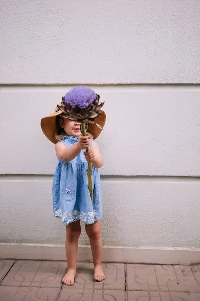 かわいい2歳の少女は 家の壁に立って屋外の大きなアーティチョークの花を保持するわら帽子をかわいい 幸せな子供時代 夏のアクティビティ ストック画像