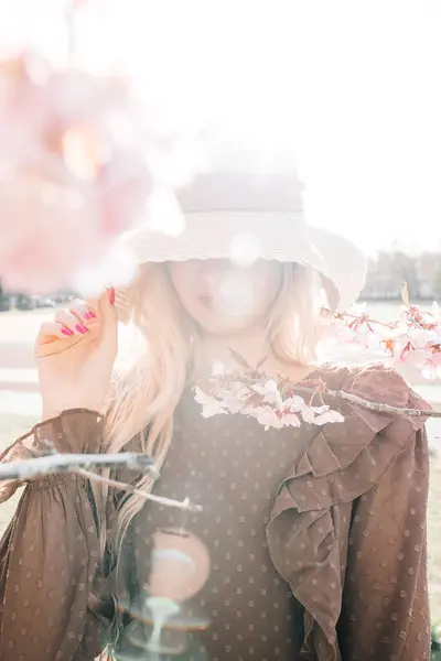 若いセンセーショナルかわいいブロンドの女性の肖像画は 咲く桜の木に立っているわら帽子の下に顔を隠し 美しいピンクのマニキュア ストックフォト