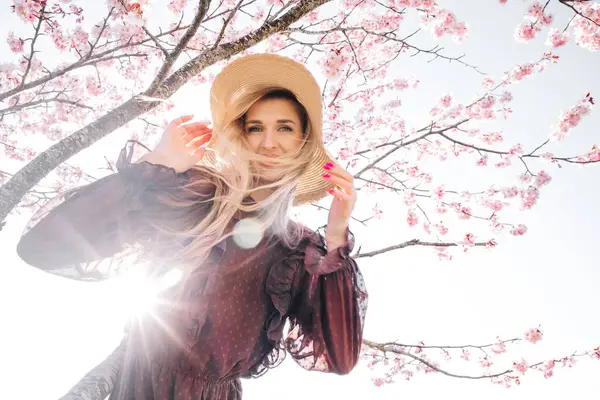 若いセンセーショナルかわいいブロンドの女性の肖像画 花開く桜の木に立っている帽子 ストックフォト