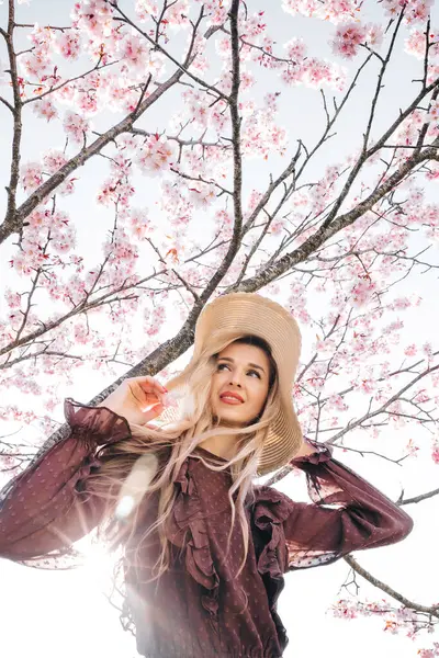 若いセンセーショナルかわいいブロンドの女性の肖像画 花開く桜の木に立っている帽子 ロイヤリティフリーのストック写真