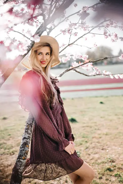 若いセンセーショナルかわいいブロンドの女性の肖像画 花開く桜の木に立っている帽子 ストック写真