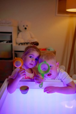 Adorable 3 years old boy and girl playing on sensory box in the kids room. Holding in hands sand sifter and looking at camera clipart