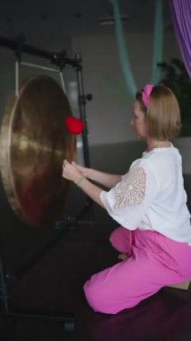 Orta yaşlı bir kadın yoga stüdyosunda büyük gongu yumuşak çekiçlerle vuruyor. İçeride ses iyileştirme terapisi. 