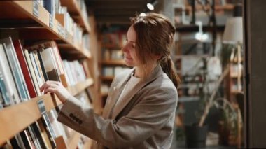 Ceketli genç mutlu kadın kitapçıda ya da kütüphanede kitap raflarında geziniyor. Kitaplara bakıyor ve hangisini seçeceğine karar veriyor gibi görünüyor..