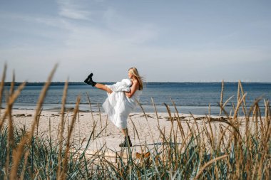 Beyaz battaniyeye sarılı genç bir kadın sahilde neşe içinde dans ediyor, sakin deniz dalgalarının altında güneşli öğleden sonranın tadını çıkarıyor..