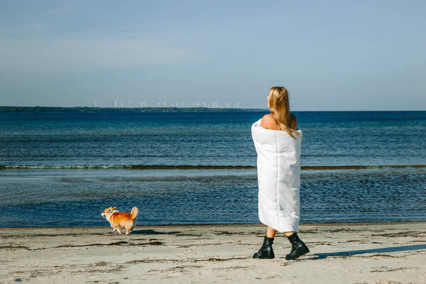 Женщина, завернутая в белое одеяло, прогуливается по пляжу с корги, наслаждаясь солнечным днем у воды возле ветряных турбин.