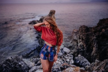 Bir kadın düşünceli bir şekilde kayalık bir plajda duruyor. Güneş arkasında sakin okyanus suyunun üzerinde batıyor..