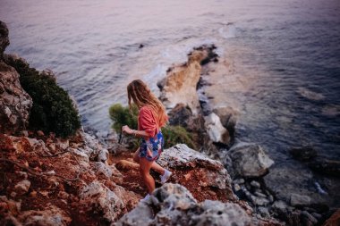 Bir kadın gün batımında yemyeşil ve okyanusla çevrili kıyı boyunca kayalık bir yolda dikkatlice yürür..