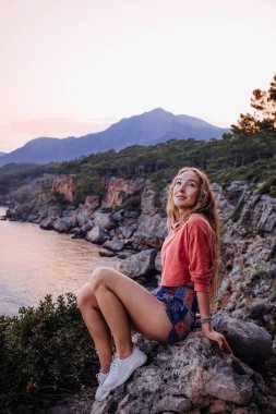 Genç bir kadın, deniz kenarındaki bir kayada dinleniyor. Kıyı şeridi boyunca gün batımının ve dağ manzarasının tadını çıkarıyor..