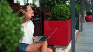 Bir kadın ahşap basamaklarda oturuyor, laptopuna odaklanmış, bir kafede canlı yeşil bitkilerle çevrili..