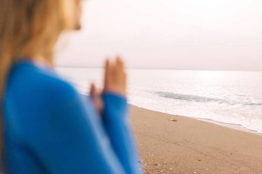 Spor elbiseli genç kadın deniz kenarında meditasyon yapıyor, sabahları plajda sakinleştirici ve topraklama egzersizleri yapıyor. Arka tarafa odaklan.